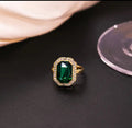Gold ring with a large green rectangular gemstone and clear crystal border on a black surface