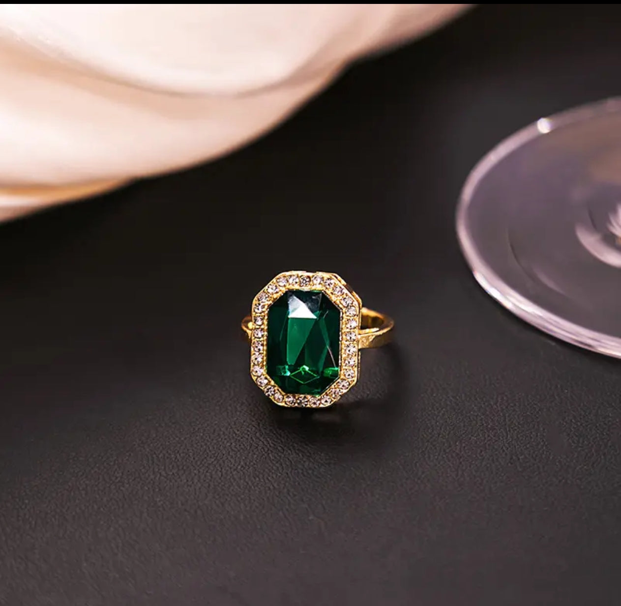 Gold ring with a large green rectangular gemstone and clear crystal border on a black surface