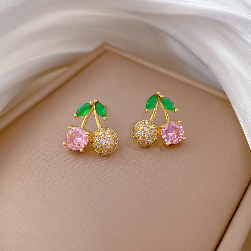 Pair of gold-tone cherry earrings with pink and clear crystals and green leaves on beige background